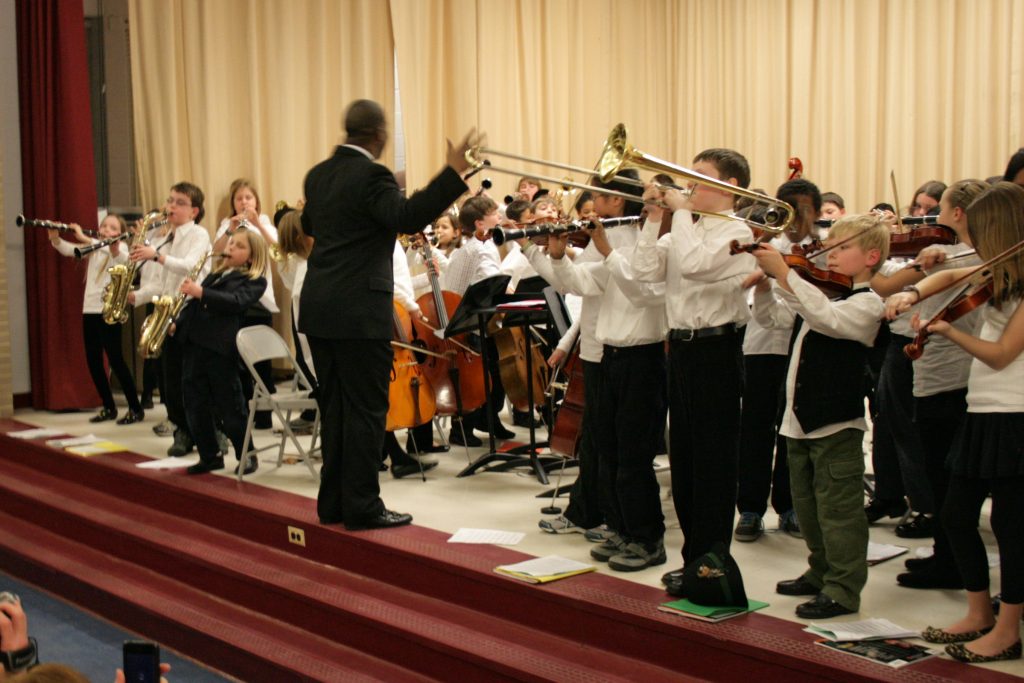 Children on stage playing various musical instruments and being directed by a man in a suit, facing them.