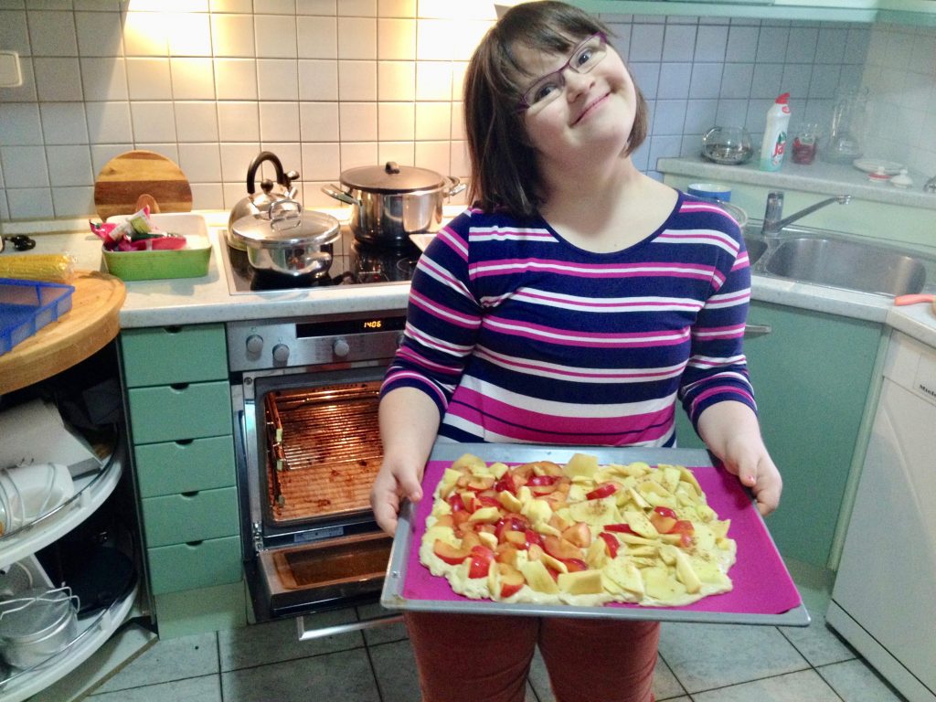 A girl with Down syndrome smiling and standing in front of an oven holding a baking tray