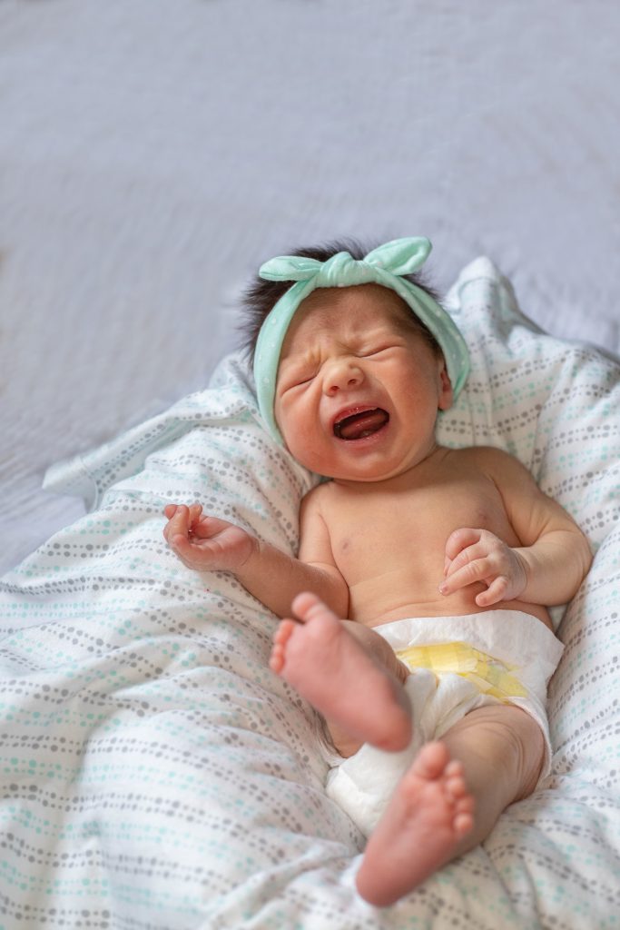 A photograph of a small baby wearing a green headband and diaper, laying on a blanket andcrying
