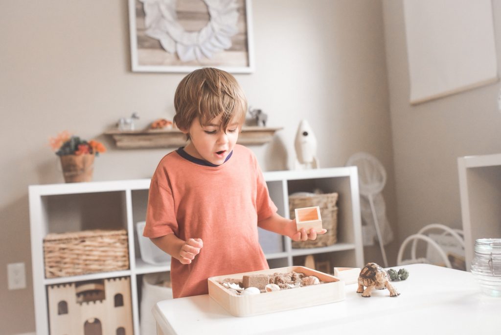 A child standing at a table with a tray of blocks and other small objects engaged in pretend play