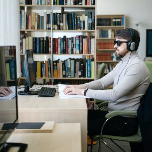 A light-skinned person with short dark hair and a beard, seated at a library computer desk, wearing dark glasses and headphones, reading from a braille book.