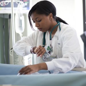 A young, dark-skinned nursing student leans over a bed while checking their watch during a simulation exercise. They have long, black hair in a ponytail, and wear white professional clothing and a stethoscope around their neck.