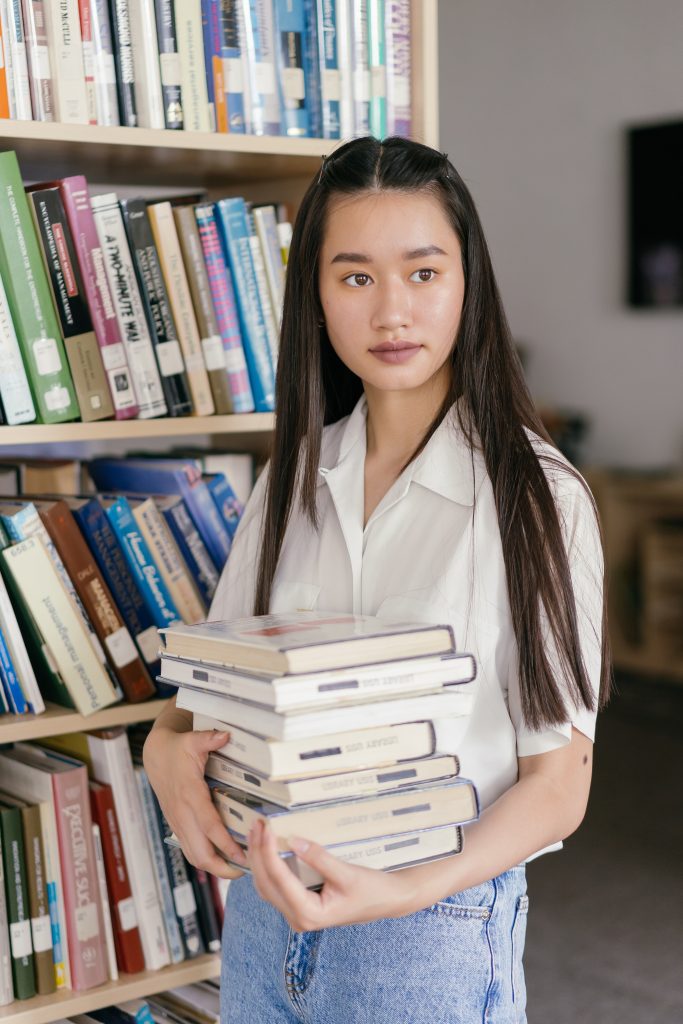 A woman stands in a library holding a stack of books.