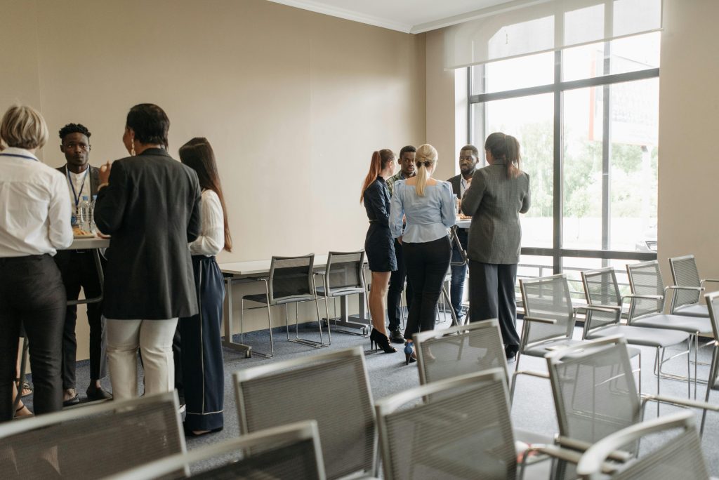 A group of professionals engaged in a discussion near a large window in a well-lit room with rows of empty chairs.