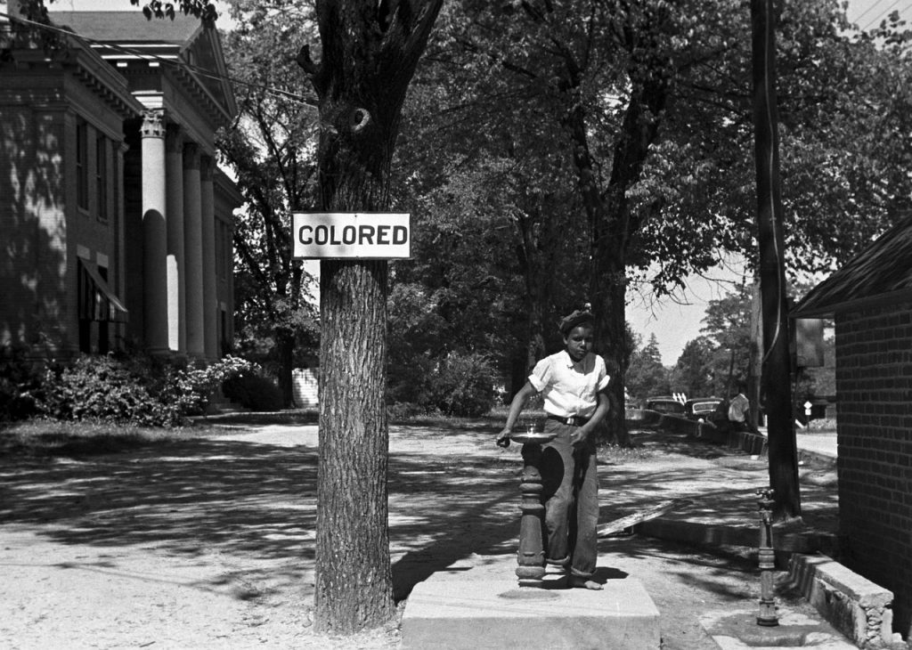 Historical black and white photo of a person drinking from a ‘COLORED’ water fountain.