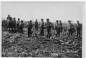 Black and white photograph of 9 young Indigenous children working in a field. Two men are in the background on horses. Text at bottom reads: "Brandon Indian School Farm"