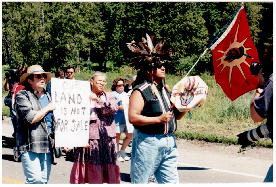 Man leads group of land defenders while holding a hand drum. Behind him are two women holding a sign that reads "our land is not for sale". In background, there are others walking alongside and a warrior flag. Flag has an image of an Onkwehon:we male with a yellow sunburst behind him.