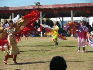Four women dance at a pow wow. Women are wearing colourful regalia dancing in the dance arena.