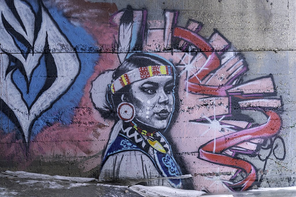Graffiti mural of an Indigenous Woman wearing regalia in Winnipeg. She is in the foreground of the mural and the background is blue and pink paint.