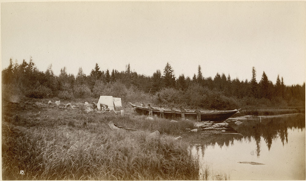 Black and white photograph of a york boat being pulled into shore. 6 people can be seen pulling york boat into shore. There is a line of pine trees in the background.