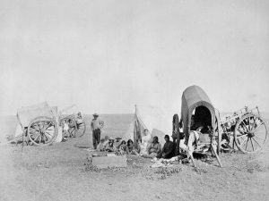 Black and white photograph of four Red River Carts and a tent. 11 people sit in the foreground of image, a mix of men, women and children.