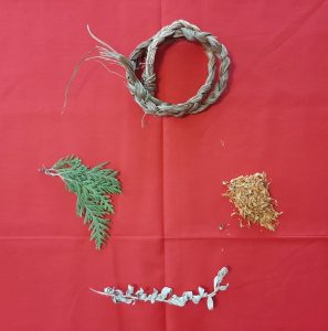 Four sacred medicines lay on red fabric in a circle. At the top lays a braid of sweetgrass, to the right lays loose tobacco, at the bottom lays a branch of white sage, and to the left lays a branch of cedar.