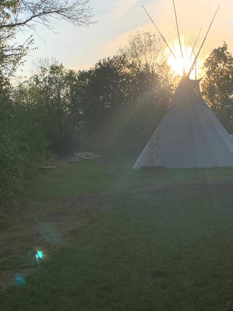 Tipi in front of a sunset. Tipi is sitting atop grass, surrounded by trees.