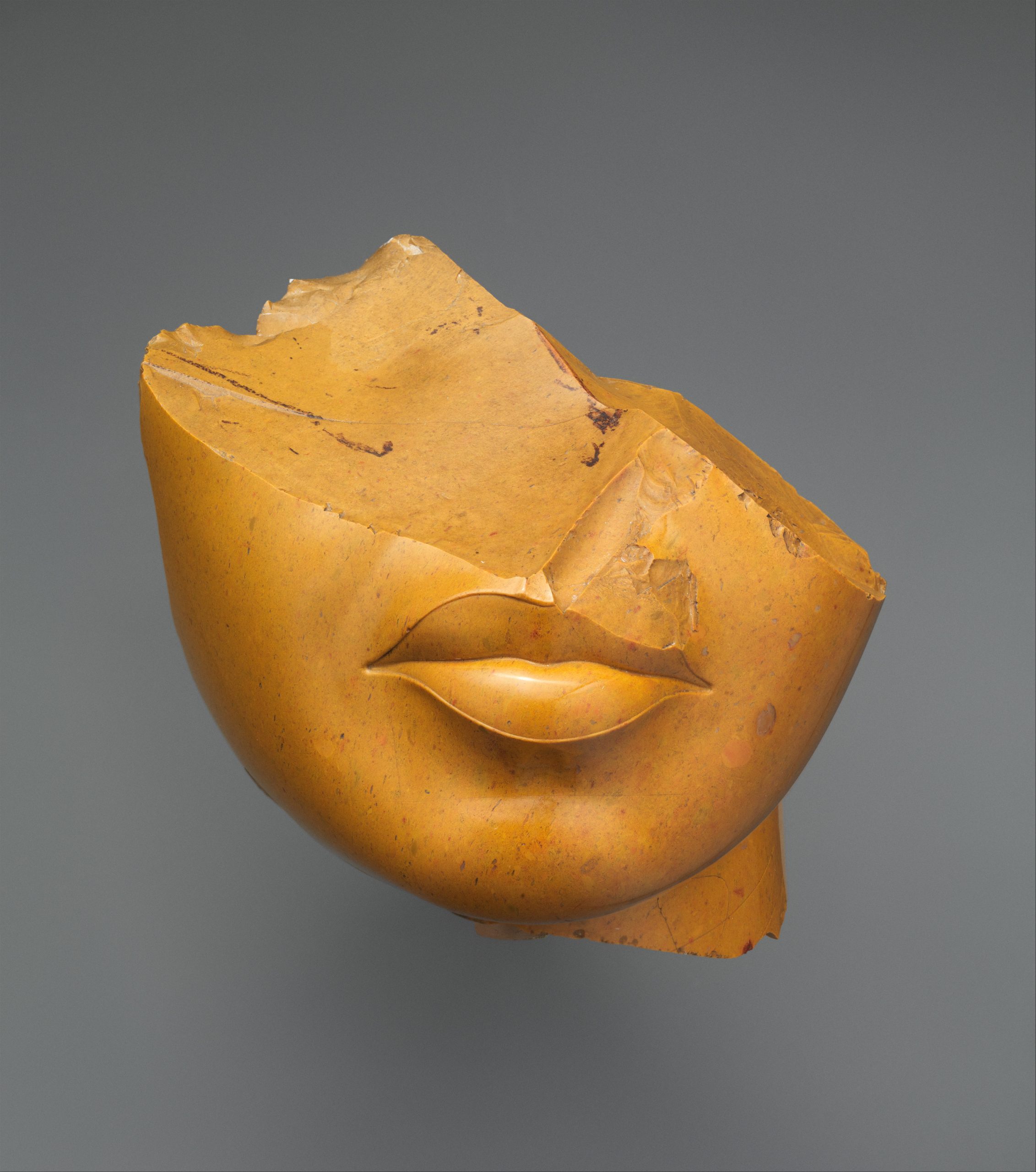 The image depicts a fragment of a statue of a Queen within the Amenhotep III or Akhenaten reign or the mid Dynasty 18. The fragment is a partial piece showing the lower half of a feminine face (i.e., the chin, lips, neck, and parts of the cranium). The medium used is yellow jasper. The dimensions of this piece are h. 13 cm (5 1/8 in); w. 12.5 cm (4 15/16 in); d. 12.5 cm (4 15/16 in). The piece is housed within the Metropolitan Museum.