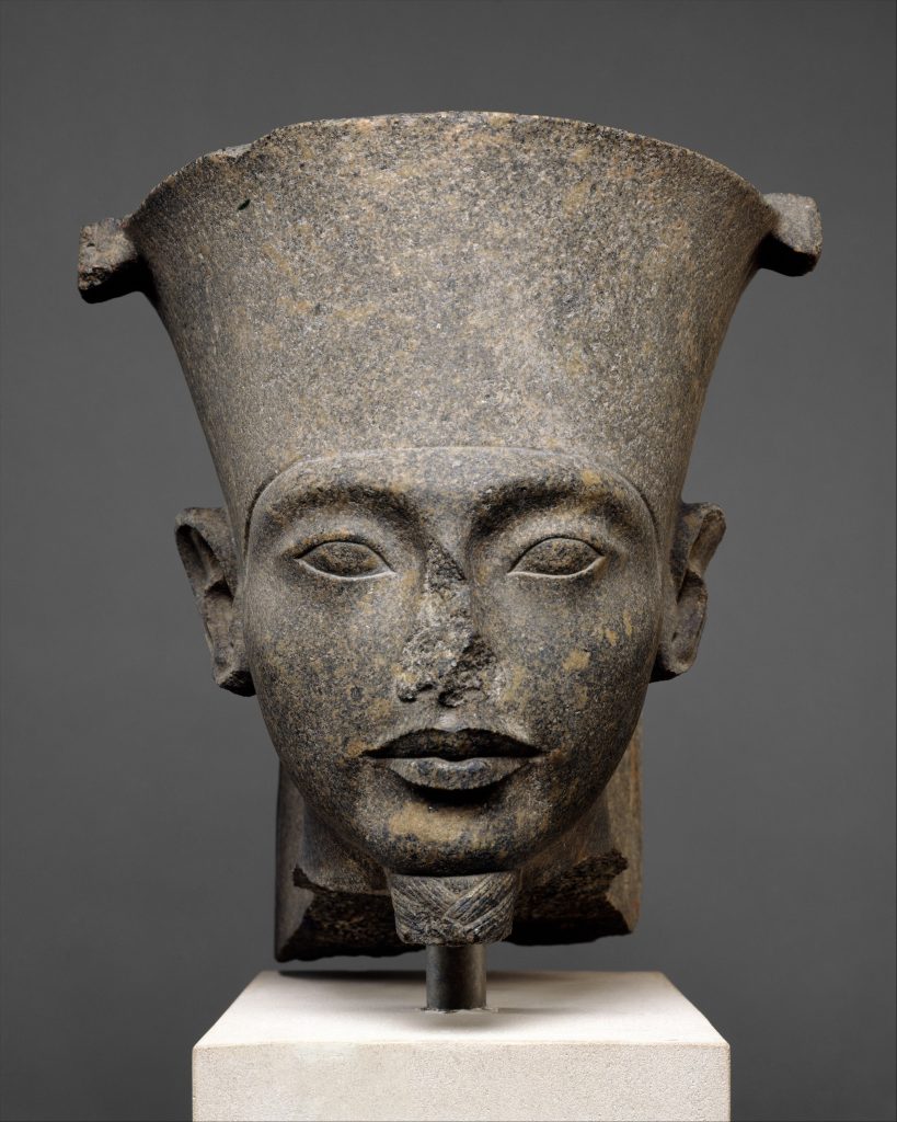 A stone statue of a standalone head missing a nose. The head is adorned with a tall flat hat. The face also has a chin adornment that is rectangular in shape that fits the chin.