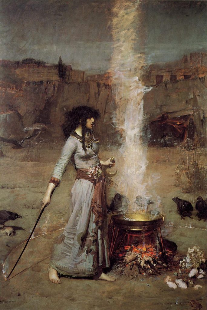 A tall, slender woman, with brown curly hair standing over a fire pit holding a sword. The woman is wearing a long ankle length gown outfitted with a belt, neck, and wrist cuffs. The background is a rocky desert scene.
