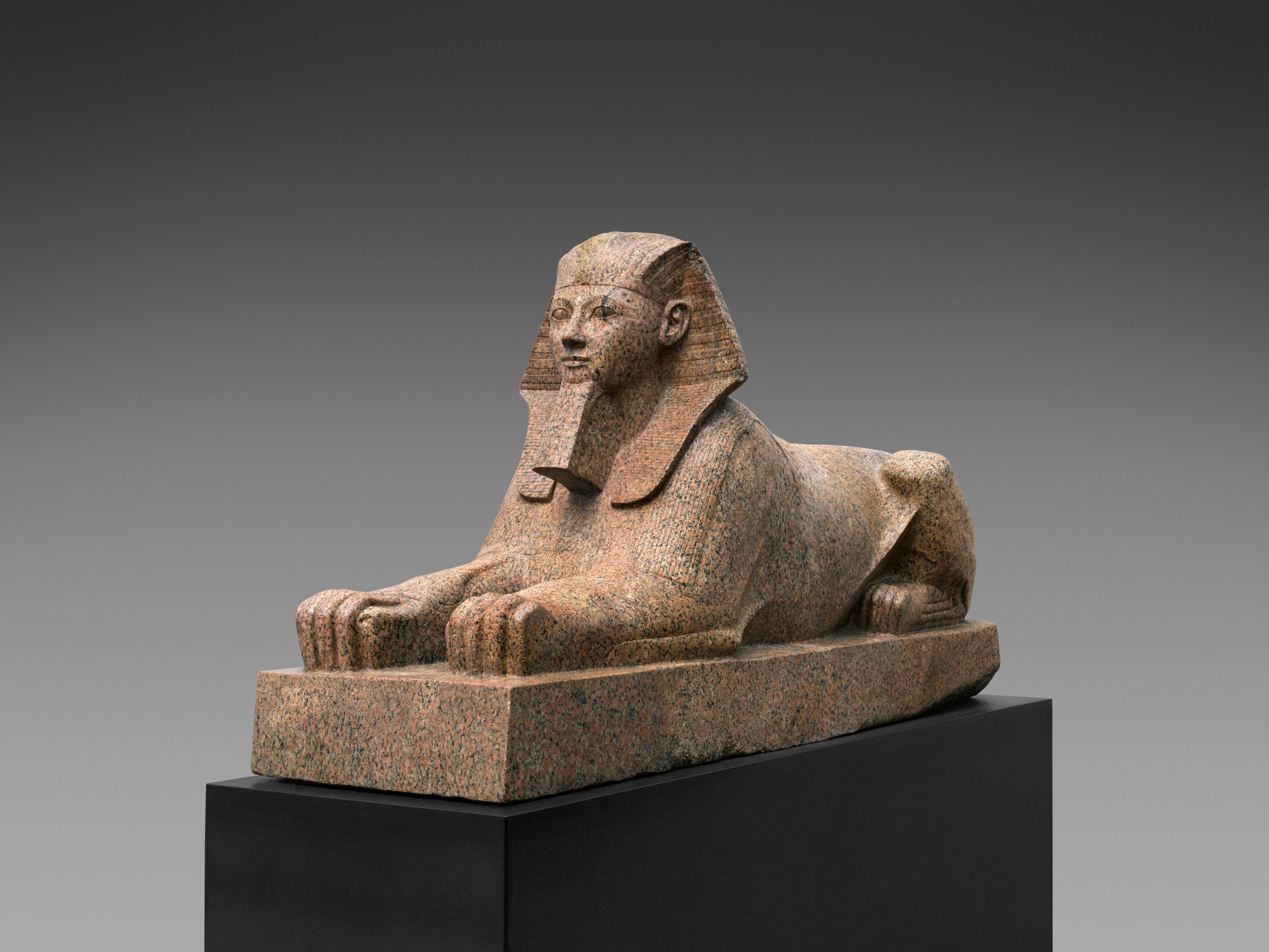A statue of a Spinx. The Sphinx is beige in colour and rests laying prone on a piece of slab.
