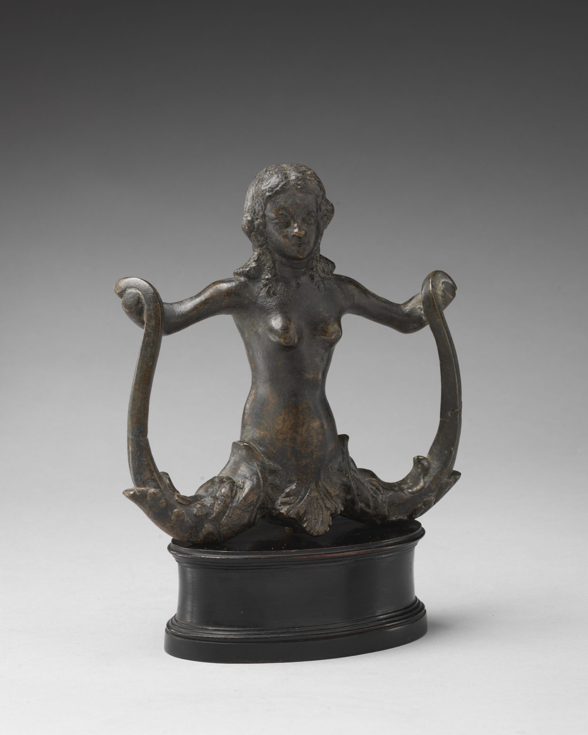 A bronze sculpture of a mermaid holding its bisected tail in each hand with arms outstretched on a pedestal