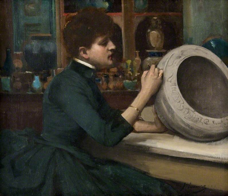 A woman sits in a room full of pottery while she uses her right hand to detail an ornate piece of pottery.