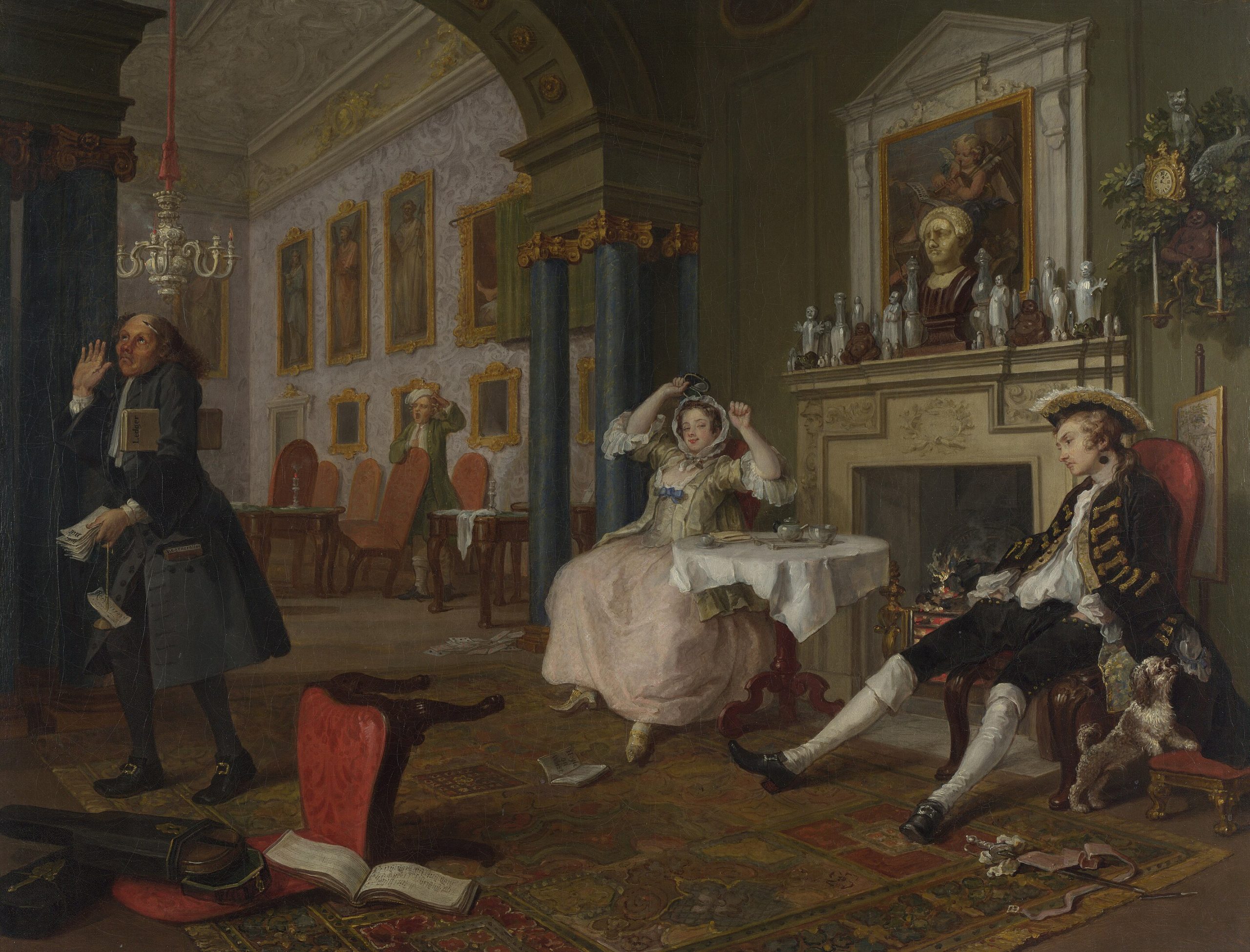 A seated man and woman in a room watching a man whose back is turned towards them shrug his shoulders beside a fallen chair