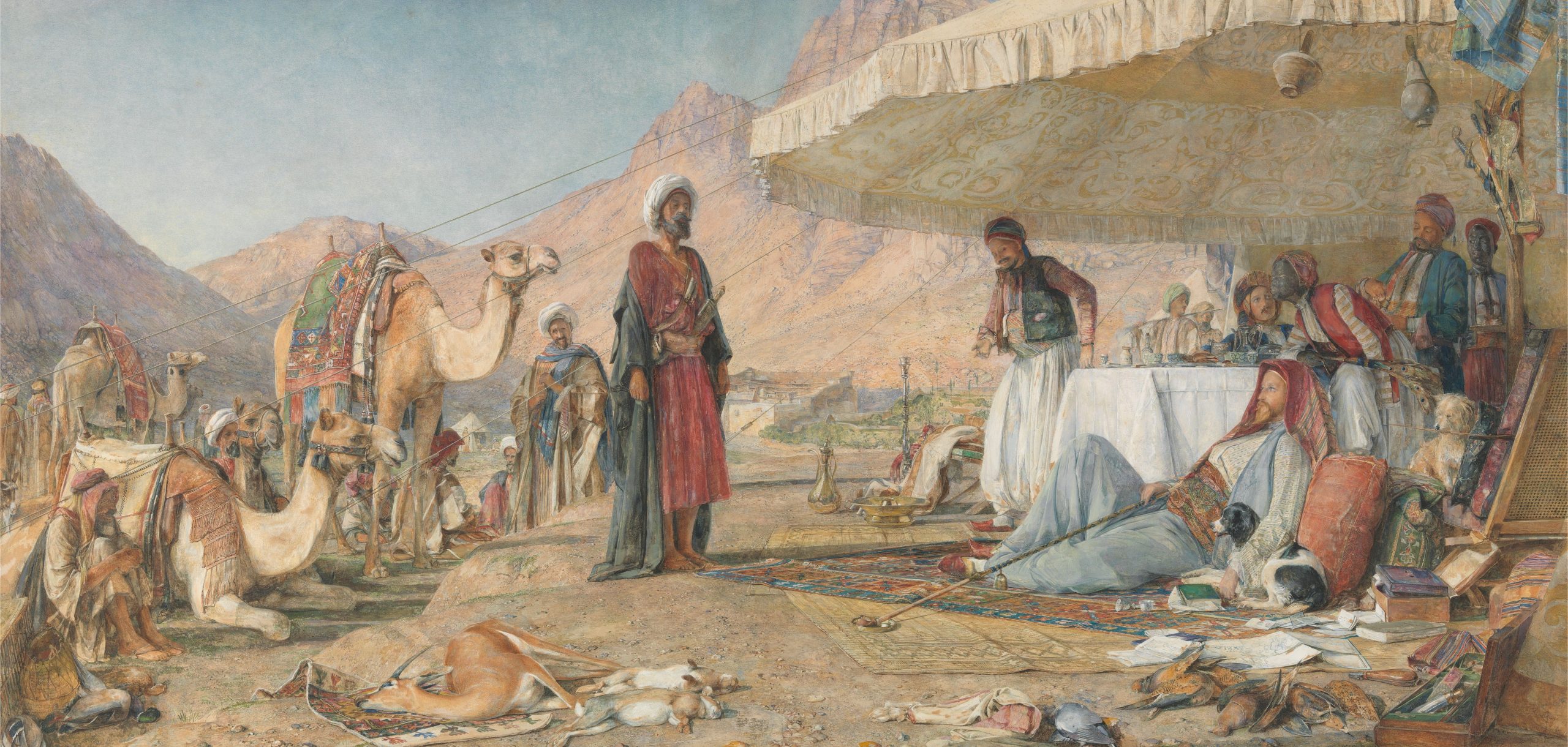 A man stands to confront another man who lies on the ground surrounded by civilians and camels in a mountainous landscape.