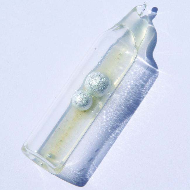 A sealed, tube-like glass container is shown. The container is partially filled with a colorless liquid and contains two metallic spheres.